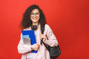 young curly student woman wearing backpack glasses holding books and tablet over isolated red backgr