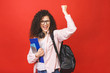 © denis_vermenko - Happy winner celebrating victory. Young curly student woman wearing backpack glasses holding books and tablet over isolated red background. Pointing finger.
