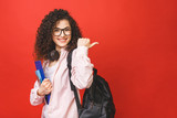 Fototapeta  - Young curly student woman wearing backpack glasses holding books and tablet over isolated red background. Pointing finger.