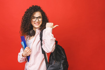 Young curly student woman wearing backpack glasses holding books and tablet over isolated red background. Pointing finger.