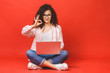 © denis_vermenko - Happy young curly beautiful woman sitting on the floor with crossed legs and using laptop on red background. Ok sign.