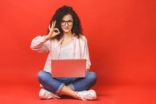 Happy Young Curly Beautiful Woman Sitting On The Floor With Crossed Legs And Using Laptop On Red Background. Ok Sign.