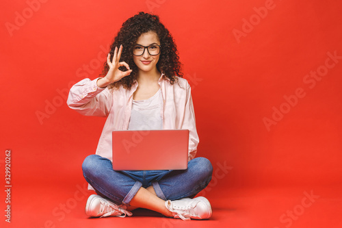 Fotomural Happy young curly beautiful woman sitting on the floor with crossed legs and using laptop on red background