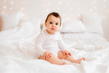 Wall Mural - healthy baby boy 10 months old in white clothes smiling sitting on white bed linen in bed, space for text