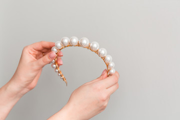 Close up of hands showing a trendy bridal gold diadem (hair hood, hair band, tiara) with big pearls. Wedding accessories. Copy space. Gray background.