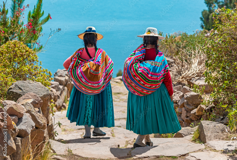 Two indigenous Quechua women in traditional clothes walking down the path to the harbor of Isla Taquile (Taquile Island) with the Titicaca Lake in the background, Peru. - obrazy, fototapety, plakaty 