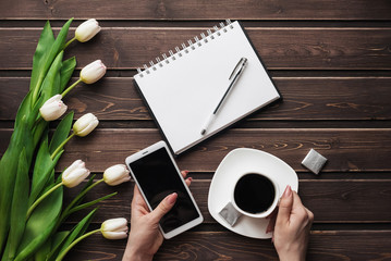 Wall Mural - White tulips on a wooden table with an empty notebook, smartphone and a Cup of coffee in women's hands