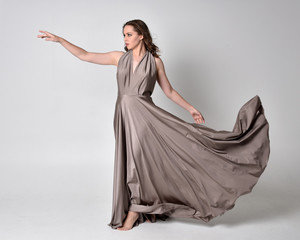 Poster -  Portrait of a pretty brunette girl wearing a long silver evening gown, full length standing pose against a studio background.