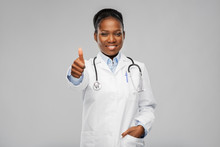 Medicine, Profession And Healthcare Concept - Smiling African American Female Doctor In White Coat With Stethoscope Showing Thumbs Up Over Grey Background