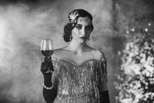 Black And White Closeup Portrait Stylish Retro Woman In Shine Silver Dress With Glass Of Wine In Hand. Old Finger Wave Hairstyle With Headband. Vintage Holiday. Backdrop Bright Smoke. Cat Eyes Makeup