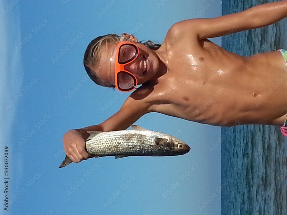 Portrait Of Happy Shirtless Girl Holding Dead Fish By Sea Against Sky - obrazy, fototapety, plakaty 