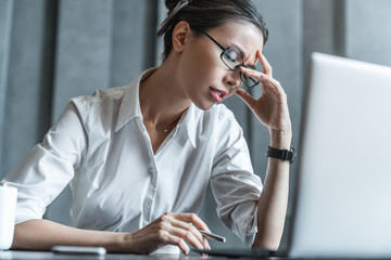 Stressed young female office worker sitting at desk holding head because of pain in office