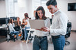 © standret - Man and woman in formal wear standing with laptop in hands indoors in office with young people that works behind them