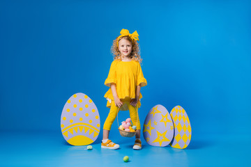 Wall Mural - child a blonde girl in a yellow dress is smiling standing with a row of large decorative Easter eggs on a blue background