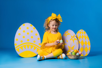 Wall Mural - child a blonde girl in a yellow dress is smiling standing with a row of large decorative Easter eggs on a blue background