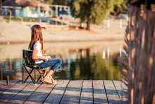 Little Cute Girl Sitting On A Wooden Pier With Fishing Rod.