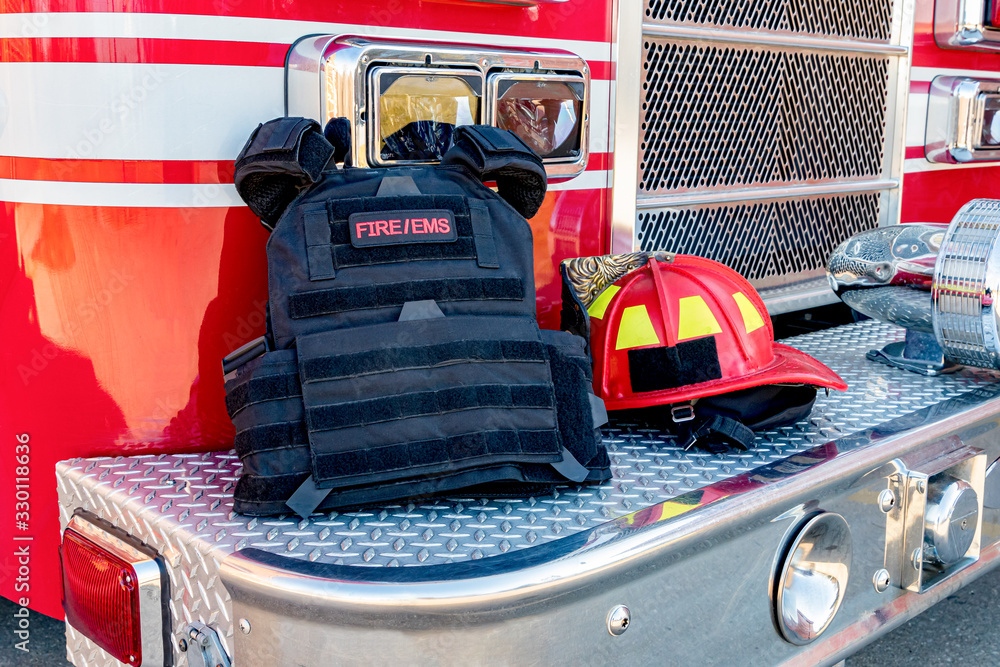 Ballistic vest and red firefighter helment on fire truck bumper. Concept of evolving role of fire department response to mass casualty shooting and terrorism - obrazy, fototapety, plakaty 