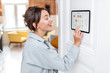 © rh2010 - Young woman controlling home with a digital touch screen panel installed on the wall in the living room. Concept of a smart home and mobile application for managing smart devices at home