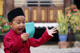 Fototapeta Las - A Malay boy in Malay traditional cloth showing his happy reaction after receiving money pocket or 'Duit Raya' during Eid Fitri or Hari Raya celebration.