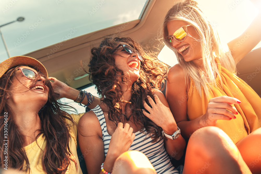 Three female friends enjoying traveling in the car. Sitting in rear seat and having fun on a road trip. - obrazy, fototapety, plakaty 