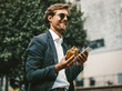 © Svyatoslav Lypynskyy - Young white colar or freelancer having a lunch on the go. Handsome man in a blue sute and sunglasses smiling holding smartphone and hamburger in his hands.