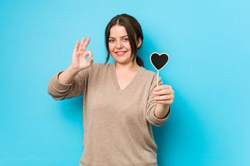 Wall Mural - Young plus size curvy woman holding a heart shape cheerful and confident showing ok gesture.