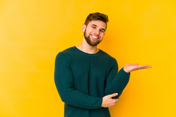 Wall Mural - Young caucasian man isolated on yellow background showing a copy space on a palm and holding another hand on waist.