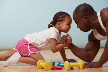 Wall Mural - Handsome black young father is arm wrestling with his cute little daughter on the floor at home. They looking at each other angrily, trying to show their resolve. Grimacing at each other.