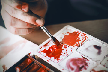 Wall Mural - The artist's dirty hand holds a paintbrush with which she mixes scarlet watercolor paint on a plastic palette. Various shades of red - scarlet, crimson, maroon.