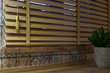Poster - Wood blinds closeup on the window, with a ribbon ladder and designer round weight, in the kitchen. Wooden windowsill and colored tiles. Indoor flower on the table.