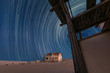 A beautiful night sky photograph with circular star trails against a deep blue sky, with an abandoned house and desert sand in the background, taken in the ghost town of Kolmanskop, Namibia.