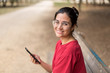 © Alberto Ortega  - Young attractive woman with glasses sitting in a bench and using her phone in a park, with a red shirt. Seville, Spain