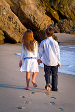 Boy And Girl Walking Hand In Hand Away From Camera On Beach Leaving Footprints In The Sand During Late Afternoon Sunlight