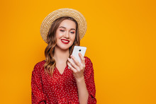Young Brunette Woman In A Red Summer Dress, A Straw Hat Chatting By Mobile Phone Isolated Over Orange Background. Online Orders, Purchases, Shopping, Credit Concept, Summer Girl With Cell