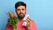 © wayhome.studio - Pollen allergy symptoms concept. Puzzled unshaven man has terrible allergy, holds plant and paper tissue, has watery eyes, looks sadly at camera, poses over blue wall, blank space. Seasonal rhinits
