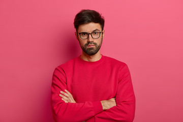 Wall Mural - Photo of self assured man with beard, keeps arms folded over body, looks seriosly at camera, wears casual clothes, has talk with colleague, poses indoor against pink background. Confident guy