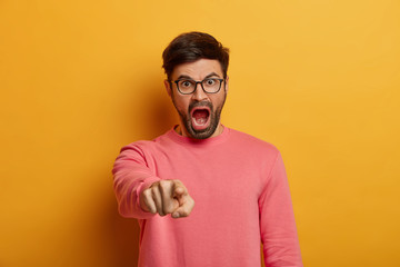 Wall Mural - Angry outraged man indicates at camera, blames you, screams from irritation, says you are guilty, wears glasses and rosy sweater, poses against yellow background, scolds person for making mistake