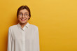 © WHstudio Leushin N - Gorgeous happy young European woman with tender toothy smile, looks aside, glad to come on formal meeting, wears round transparent glasses and white elegant shirt, poses against yellow background