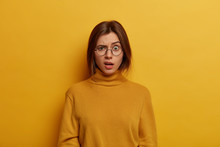 Indignant Puzzled Young Woman Raises Eyebrows And Looks With Shocked Expression At Camera, Hears Shocking Unpleasant News, Wears Round Transparent Glasses And Poloneck, Isolated On Yellow Wall