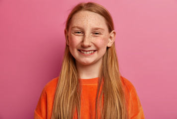 Portrait of sincere smiling girl being in good mood, grins at camera, enjoys nice day, celebrates her birthday, has long ginger hair, wears orange jumper, chuckles happily. Emotions concept.