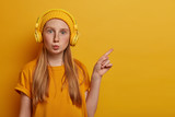 Beautiful teenager with natural ginger hair and freckles, keeps lips folded, indicates at copy space for your promo, wears stereo headphones, enjoys listening audio tracks, wears yellow clothes