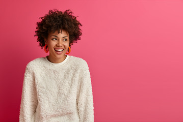 portrait of dreamy good looking woman with afro hairstyle, looks away and laughs, discusses funny re