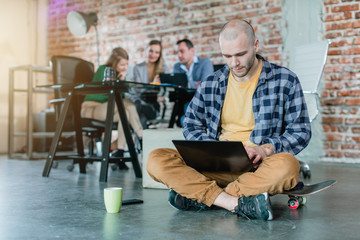 hip worker in a startup coding with laptop sitting on skateboard