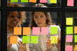 © fizkes - Diverse colleagues discussing notes on sticky papers close up, managers planning new project together, African American businesswoman writing creative ideas, tasks on sticker on glass scrum board