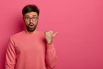 Wall Mural - Photo of stunned young bearded man indicates on right side, demonstrates something amazing, gasps with wonder, wears spectacles and casual jumper, poses against pink background. Promotion concept