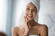 Smiling pretty young woman with towel on head looking at camera, touching cheek. Happy beautiful lady satisfied with moisturized clean skin condition, enjoying morning skincare routine in bathroom.