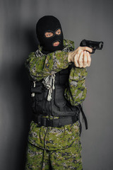 A man in camouflage uniform, body armor and a balaclava, holds his weapon ready and takes aim with a pistol, standing against a gray background.