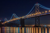 Fototapeta Nowy Jork - San Francisco Bay Bridge at night, lit up by yellow and blue lights, reflecting of the water in the Bay, long exposure