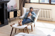 © fizkes - Smiling young man using smartphone, sitting in armchair in modern living room, happy male holding phone, looking at screen, chatting in social network or shopping online, playing game, using apps