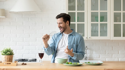 Funny young man singing song into whisk, cooking in modern kitchen, happy male holding kitchenware as microphone, listening to music, dancing, doing housework at home, preparing breakfast